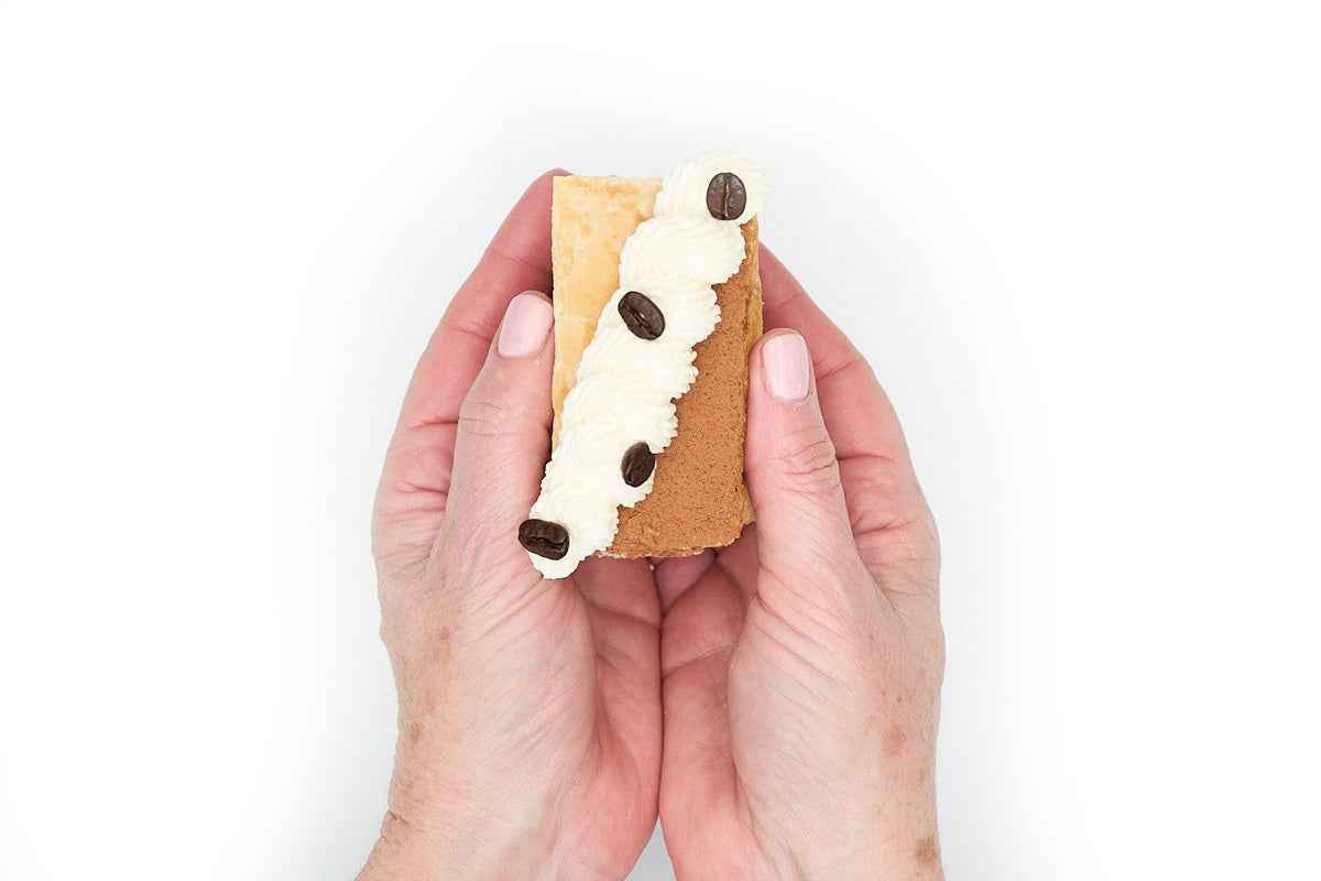 Tiramisu Napoleons held in hands, espresso pastry cream between puff layers, whipped cream and coffee bean garnish.